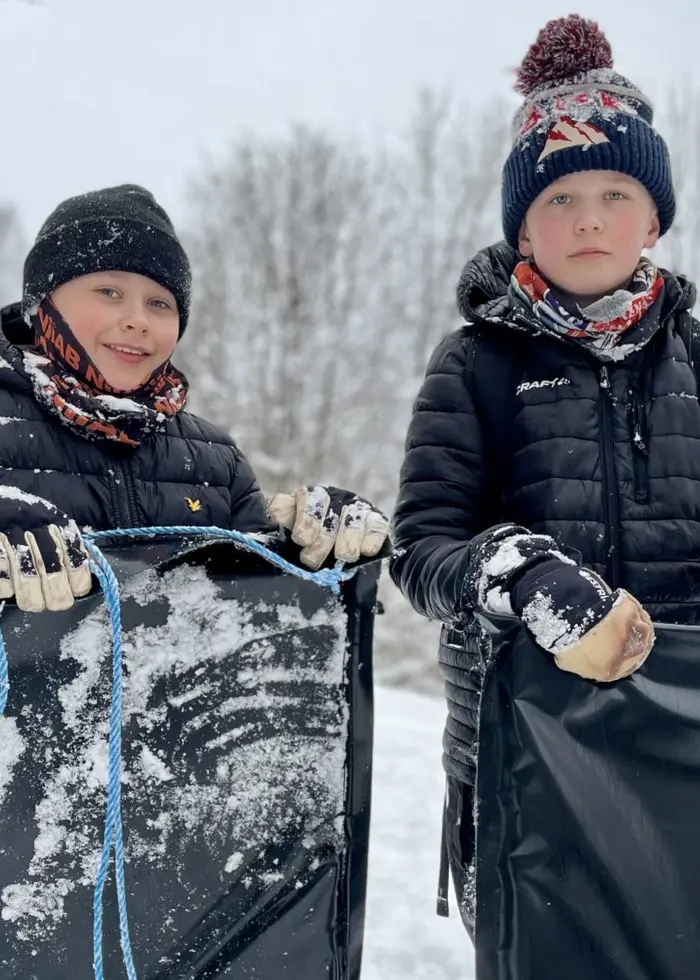 Två barn klädda i vinterkläder står utomhus i en snötäckt skog, hållande svarta madrasser.