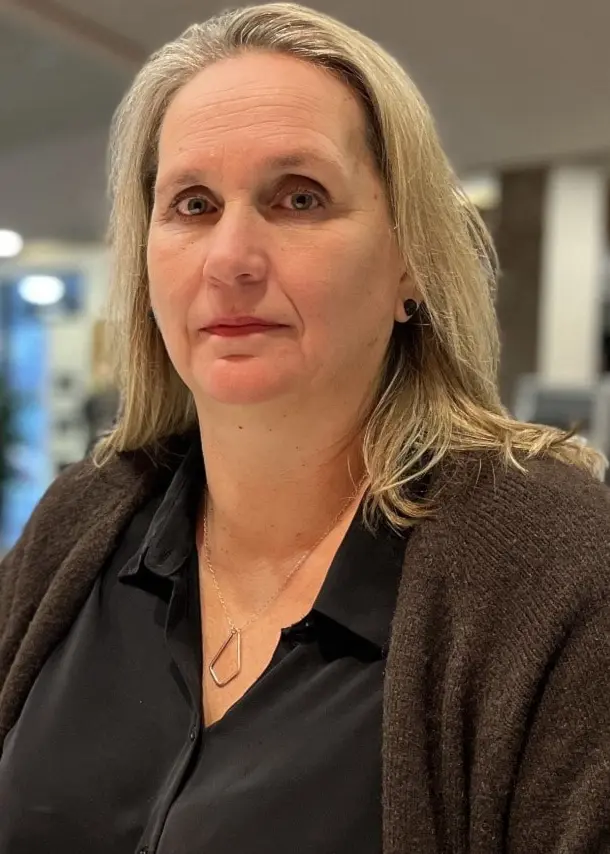 Karin Vasen står vid ett vitt bord i en modern inomhusmiljö med stora glaspartier och taklampor. I bakgrunden syns människor och möbler som skapar en arbetsplatsliknande atmosfär.