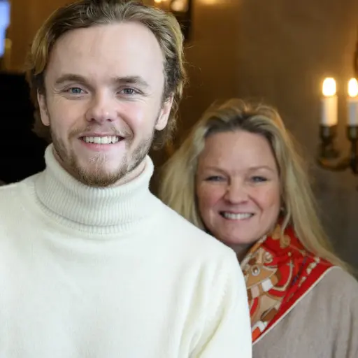 Oliver Solberg står i centrum, klädd i en vit tröja, omgiven av två personer. Bakgrunden visar en inomhusmiljö med ljuskronor och en varm belysning.