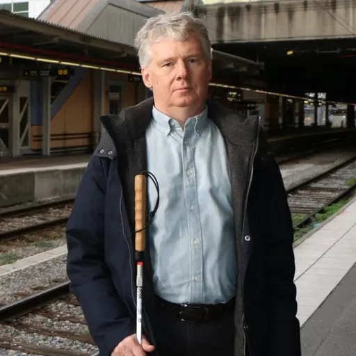 En man med vit käpp står på en tågstationsplattform. I bakgrunden syns spår, perronger och ett väntande tåg. Stationens tak och glasfasad är också synliga.