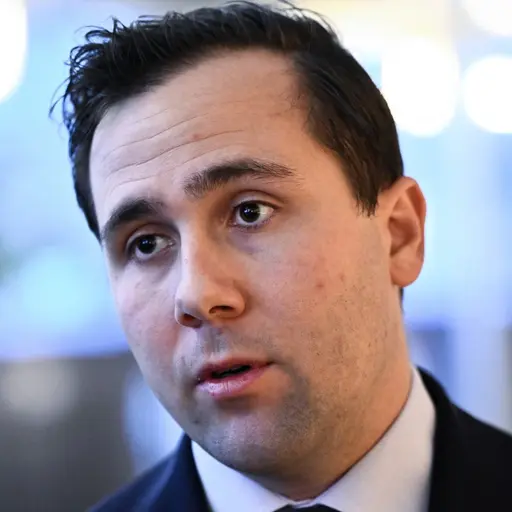 Bistånds- och utrikeshandelsminister Benjamin Dousa (M) iförd kostym, fotograferad inomhus med suddig bakgrund som visar ljuskällor och träd. Personen är i fokus.