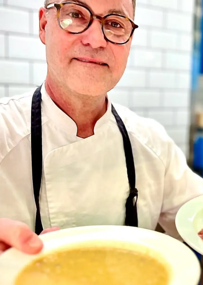 En kock i vit rock håller fram två tallrikar med mat i ett restaurangkök. På ena tallriken syns soppa och på den andra pannkakor med sylt och grädde. Bakgrunden visar vita kakelväggar och blå skåp.