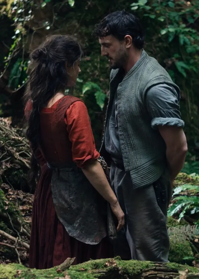 En man och en kvinna står nära varandra i en tät skogsmiljö, omgivna av gröna växter och mossa. De verkar ha en allvarlig konversation och bär avslappnade kläder i jordnära färger.