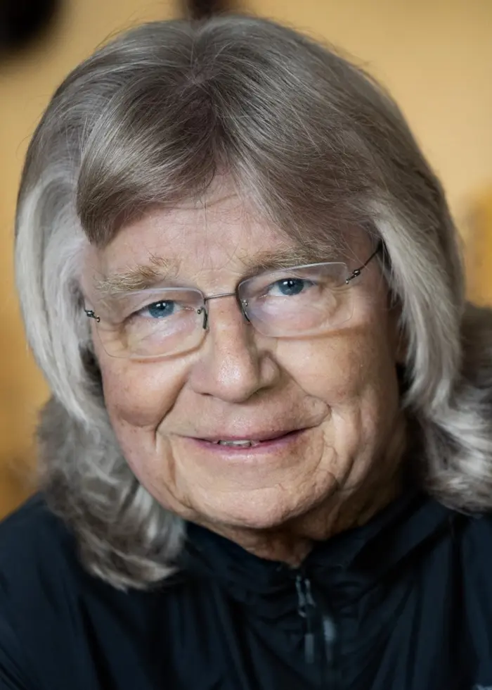 Janne Schaffer, svensk gitarrist och kompositör, sitter framför en bakgrund med upphängda gitarrer. Han har långt grått hår och bär en mörk jacka. Fokuset är på musiken och hans karriär.