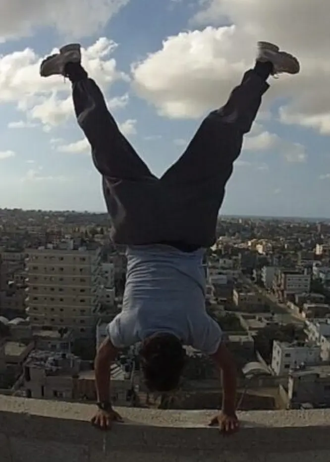 En person utför en handstående på en hög mur med en stadsvy i bakgrunden under en klar himmel med moln. Bilden förmedlar en känsla av frihet och äventyr.