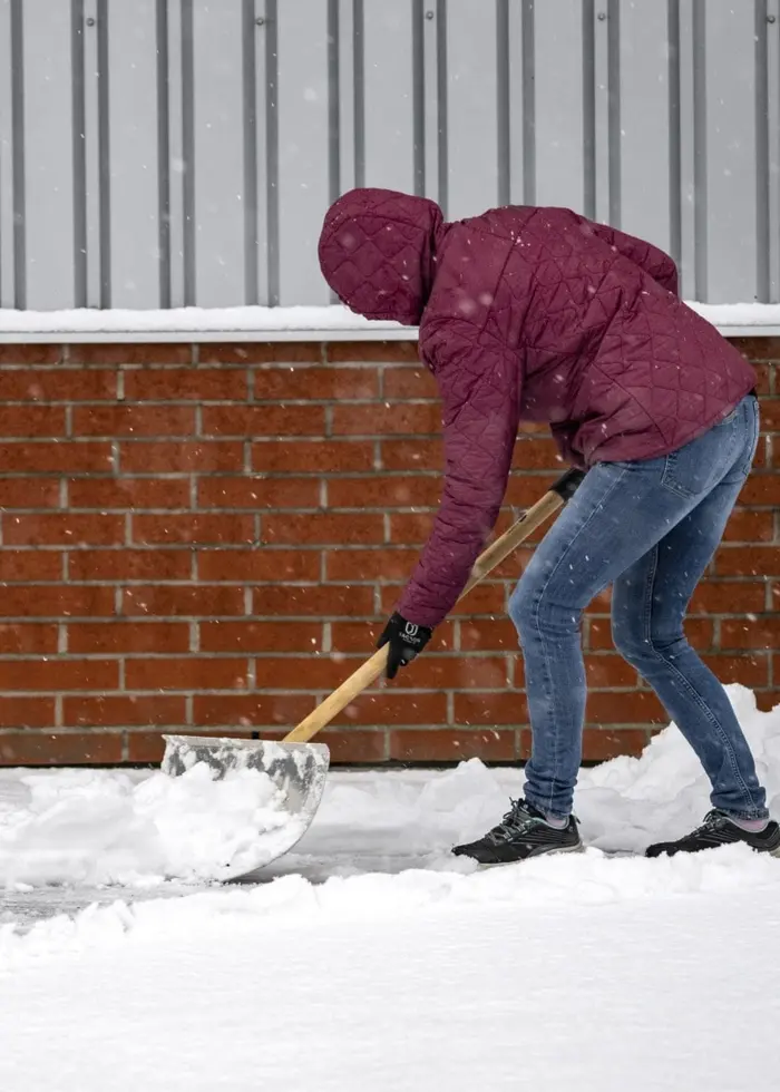 En person skottar snö framför en butik i Höör under ett snöoväder med kraftiga vindbyar. Marken är täckt av snö och en tegelvägg syns i bakgrunden. SMHI har utfärdat varningar för vädret.