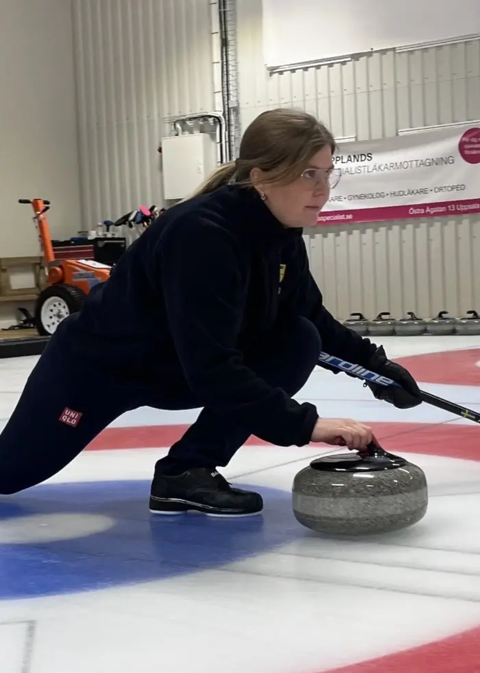 En person spelar curling inomhus på en bana med röda och blå cirklar. Personen sträcker sig framåt med en curlingsten, klädd i mörka kläder. Miljön är en träningsanläggning.