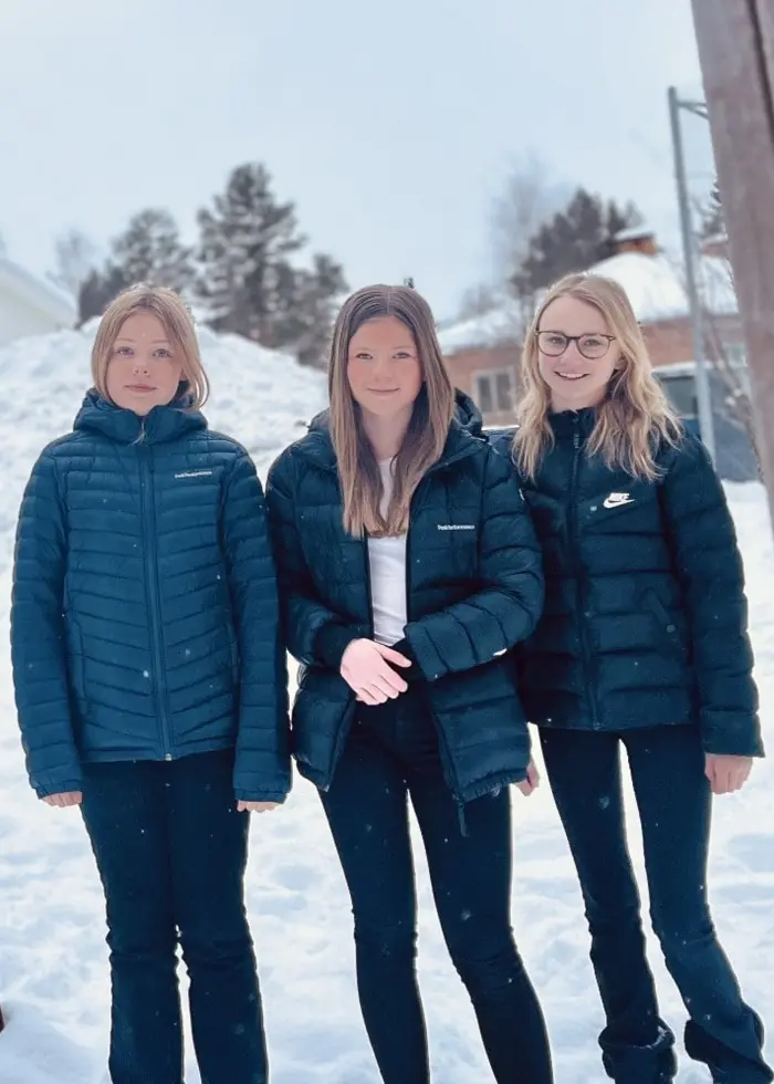 Tre unga personer, identifierade som Alva Söder, Ella Söder och Anna Larsén, står utomhus på en lekplats i vintrig miljö med snö på marken. De bär mörka jackor och ser mot kameran.