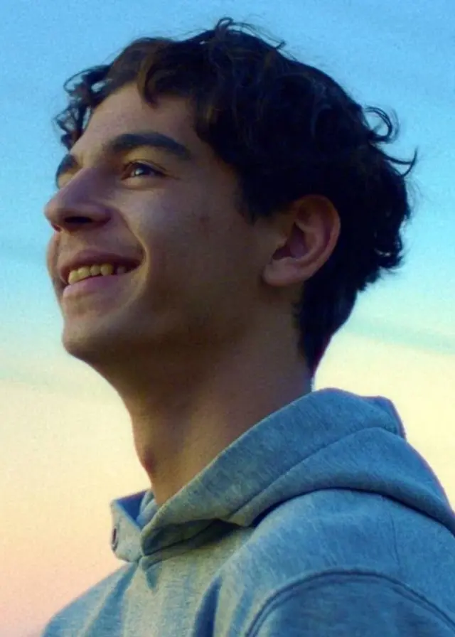 En person står utomhus i en grå huvtröja med en klar himmel och träd i bakgrunden. Solnedgången färgar himlen i mjuka nyanser av blått och rosa.