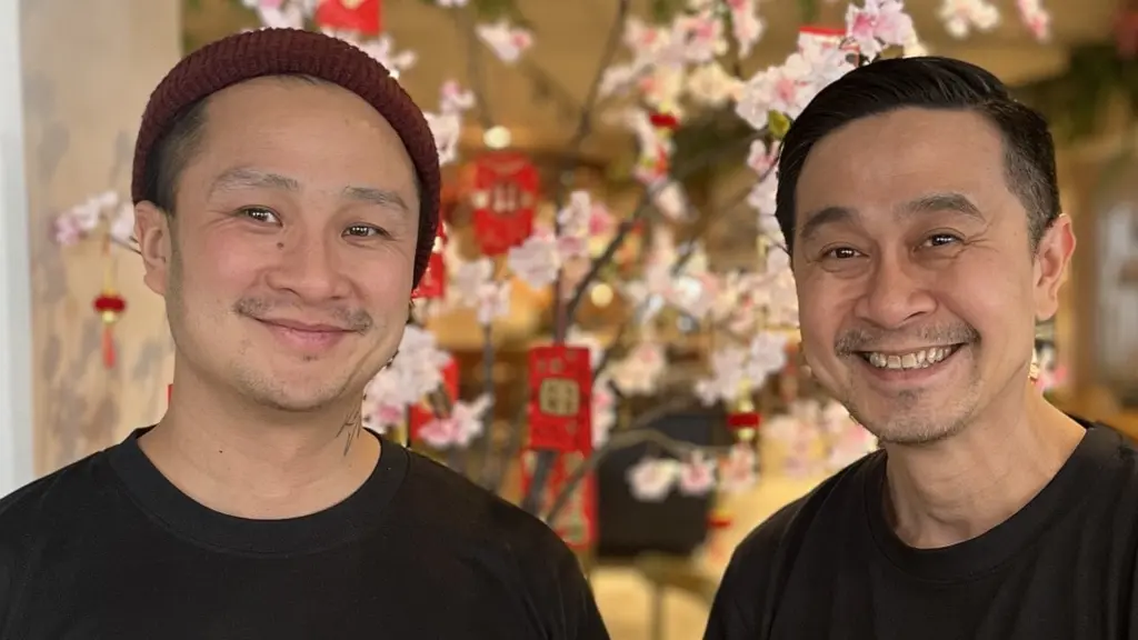 Två personer står framför ett dekorativt träd med rosa blommor i en restaurangmiljö. De bär svarta t-shirts med tryck på bröstet. I bakgrunden syns en disk och hyllor.