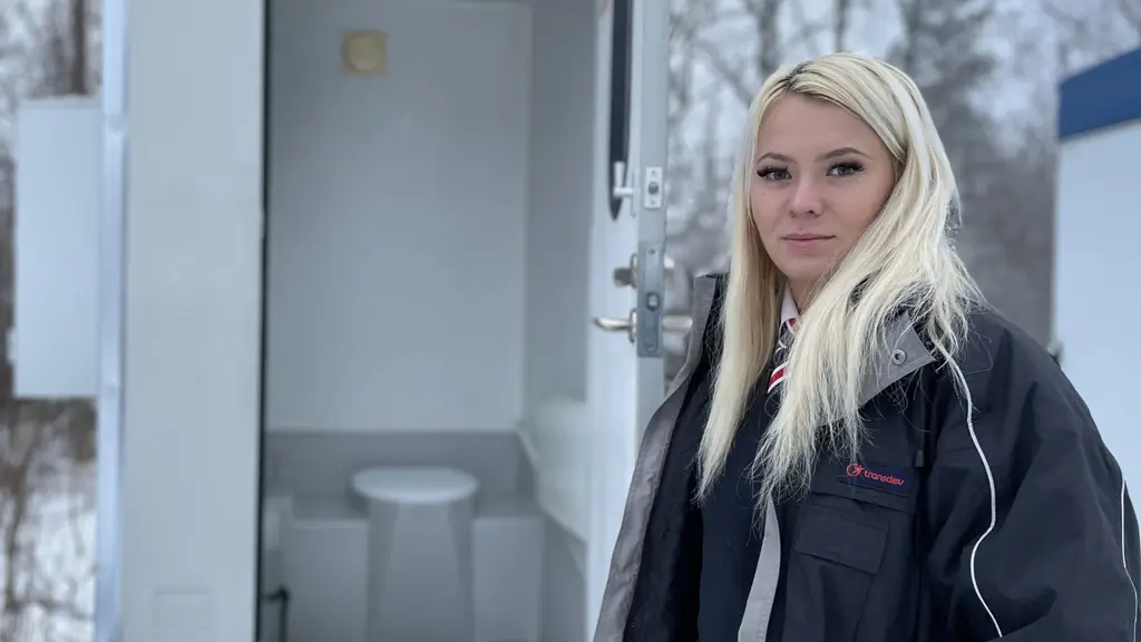 Ung kvinna i chaufförsdräkt står framför den öppna dörren till ett utedass