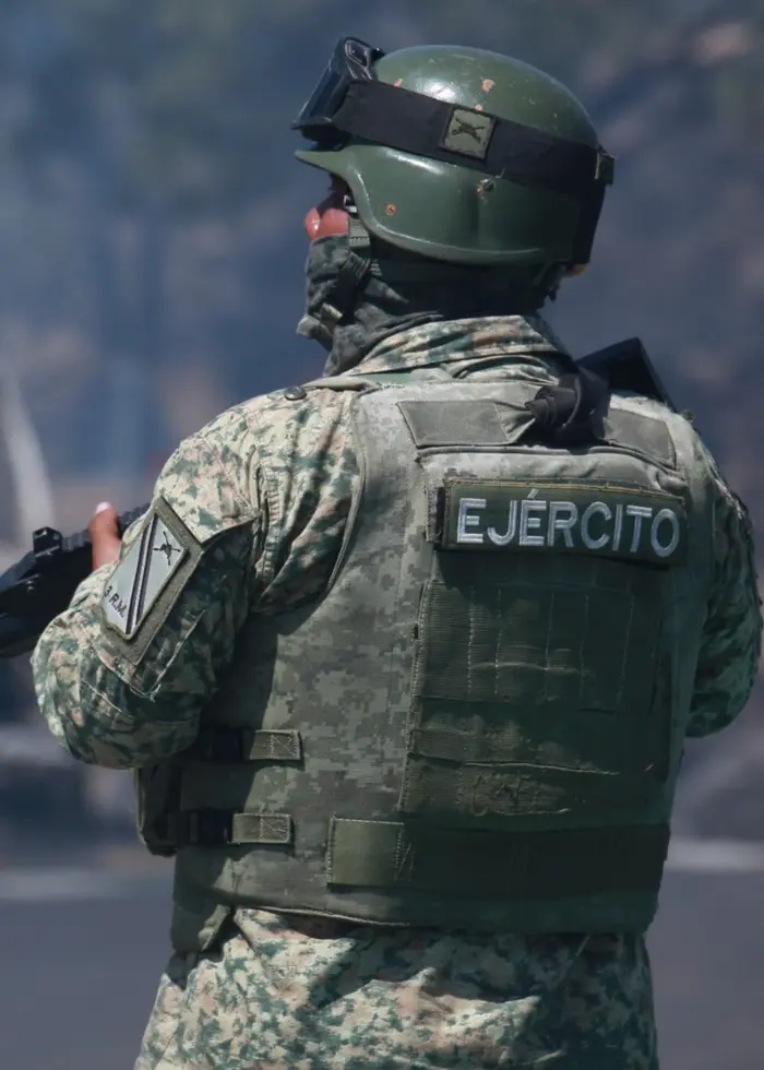 En mexikansk soldat i uniform och hjälm står med ett gevär på en väg. I bakgrunden syns ett utbränt fordon och rök som stiger upp, vilket tyder på en våldsam händelse.