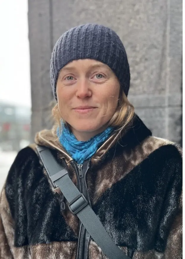 Tre personer står utomhus i en stadsmiljö med vinterkläder. Bakgrunden visar byggnader och en gata med snö. Personerna är klädda i vinterkläder.