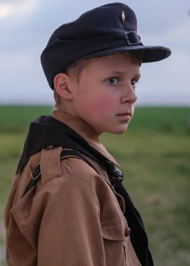 Ett barn i uniform står utomhus framför ett hus med vita väggar och stora fönster. Ett cykelstyre syns i bakgrunden, och landskapet är öppet med gröna fält.