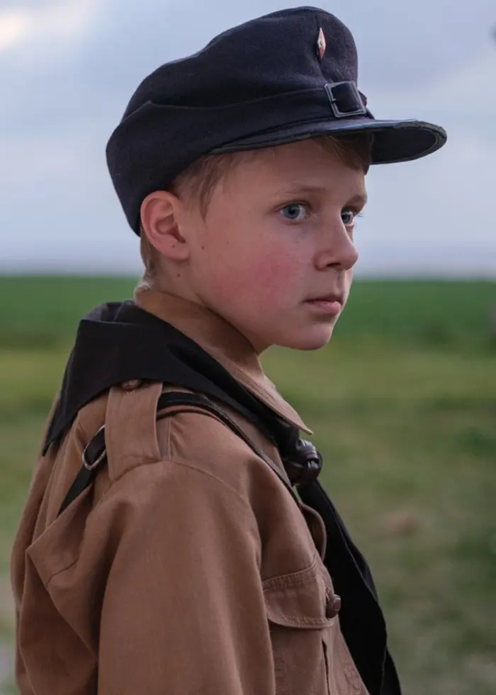 Ett barn i uniform står utomhus framför ett hus med vita väggar och stora fönster. Ett cykelstyre syns i bakgrunden, och landskapet är öppet med gröna fält.