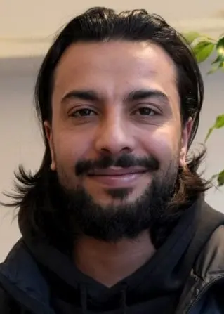 Han har på sig en svart jacka och en svart hoodie. Står framför en vit vägg och ett träd inomhus.