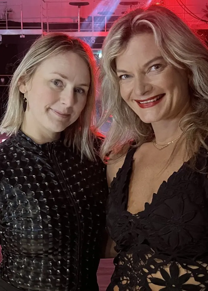 Två personer i eleganta svarta kläder står i en dansstudio med färgglada ljus och en trappa i bakgrunden. Miljön ger en känsla av en fest eller uppträdande.