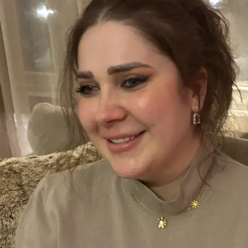 En kvinna sitter på en beige soffa med en ljusgrå tröja. Bakgrunden visar ett fönster med gardiner och svag belysning. En dekorativ kudde syns till vänster på soffan.