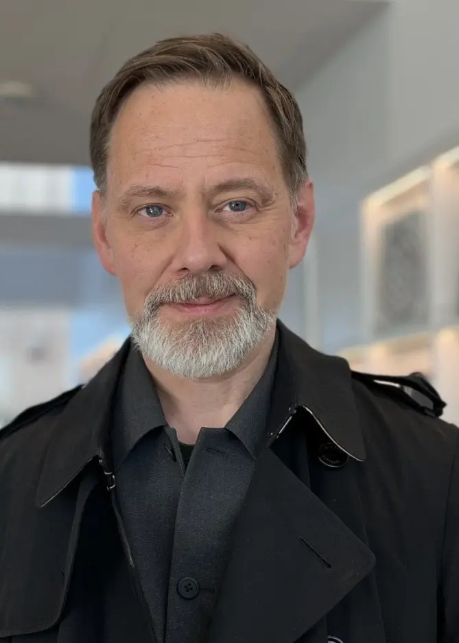 Två personer stående framför olika bakgrunder. Den vänstra personen har rödbrunt hår och en randig tröja, framför en tegelvägg. Den högra personen är klädd i svart kappa och befinner sig i en modern inomhusmiljö.