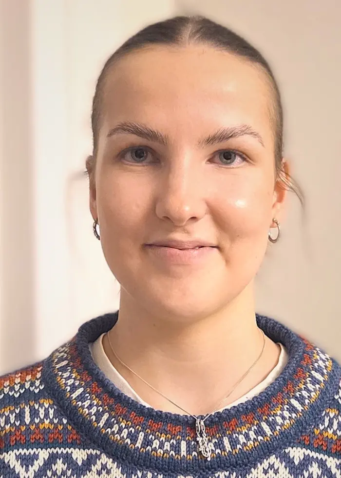 En person klädd i en stickad tröja med ett färgglatt mönster står framför en ljus vägg inomhus. Bakgrunden är enkel med en dörr synlig till vänster.