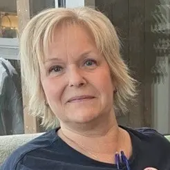 En person sitter på en grön soffa bredvid en svart hund som ligger avslappnat på en vit kudde. I bakgrunden syns en stor grön växt och en fönstervägg som släpper in dagsljus.