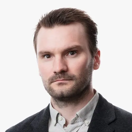 Kronprinsessan Mette-Marit syns i en mörk kavaj med långt blont hår. Bredvid är en man i mörk kavaj och skjorta. Bakgrunden är oskarp och en del av bilden är en studiofotografering.