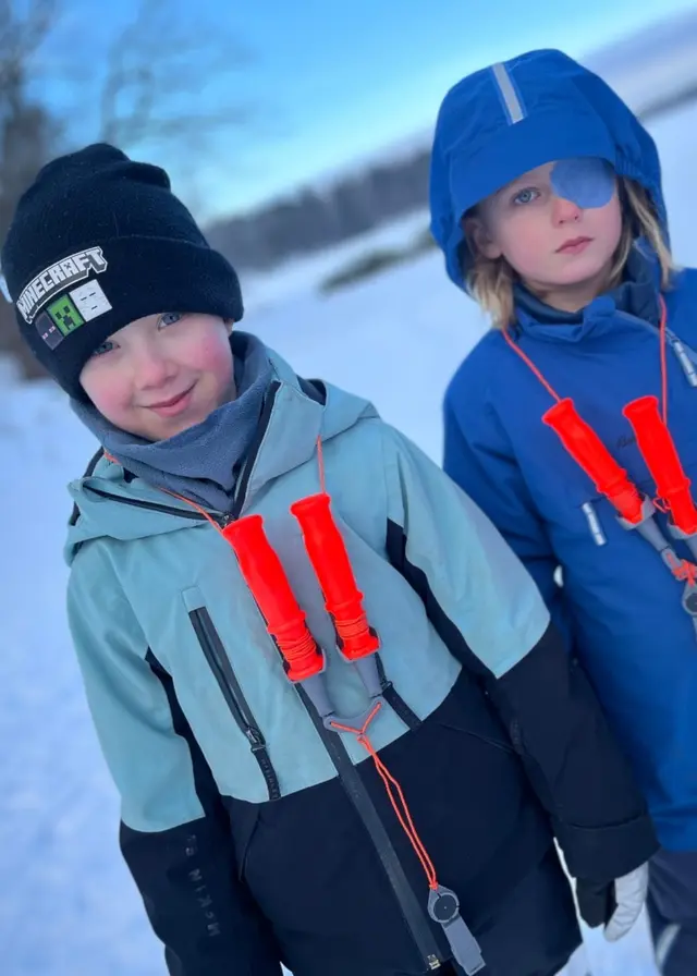 Två skolelever i vinterkläder står utomhus på snötäckt mark. Bredvid dem håller en person upp en stor gädda under en fisketur i vinterlandskap.