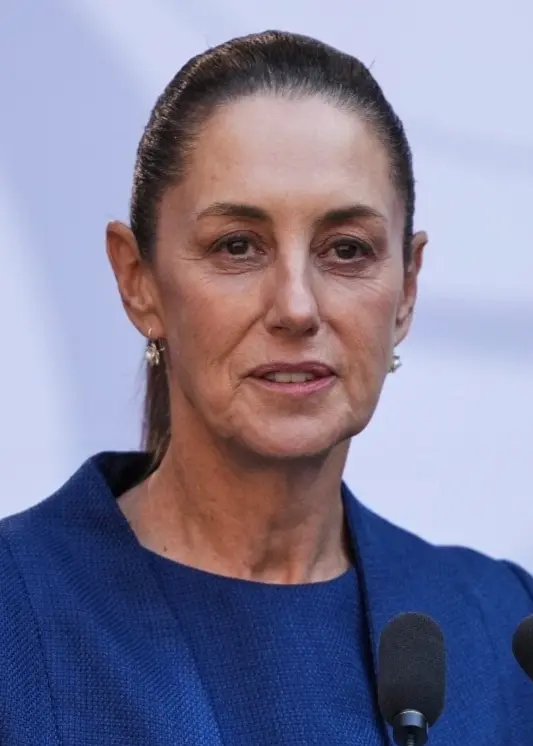 Mexikos president Claudia Sheinbaum står vid ett podium iklädd en mörkblå kavaj. Bakgrunden är ljus med subtila mönster, och framför henne finns mikrofoner.