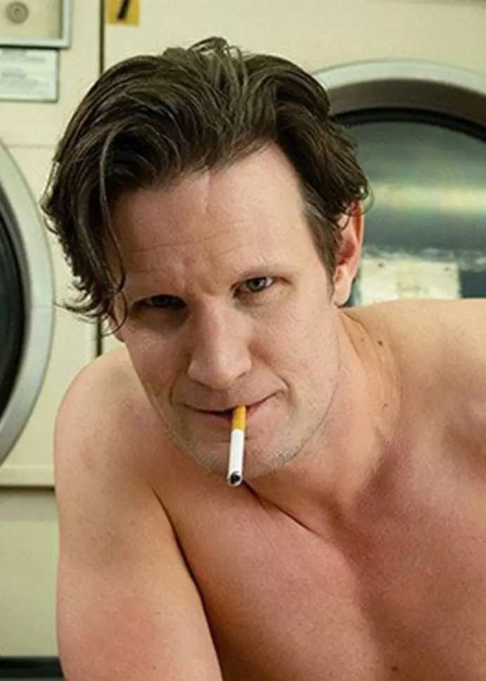 Två personer sitter i en tvättomat. Den ena är barbröstad och lutar sig framåt, medan den andra bär randiga kläder och solglasögon. Bakom dem syns stora tvättmaskiner.