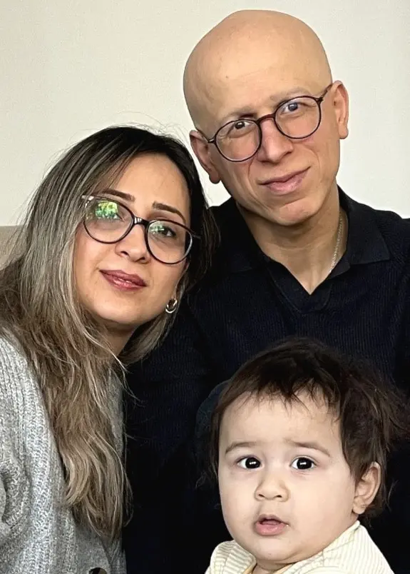 En familj sitter tillsammans i ett vardagsrum. En vuxen och ett barn leker vid en grön leksak. I bakgrunden syns en beige soffa och ett ljust trägolv.