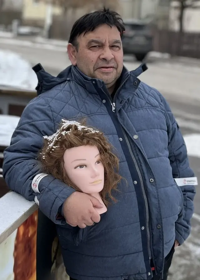 En person står vid ett bord utomhus med flera dockhuvuden med peruker. Bordet är täckt av ett tunt lager snö och en svensk flagga syns delvis. I bakgrunden ses en snötäckt gata.