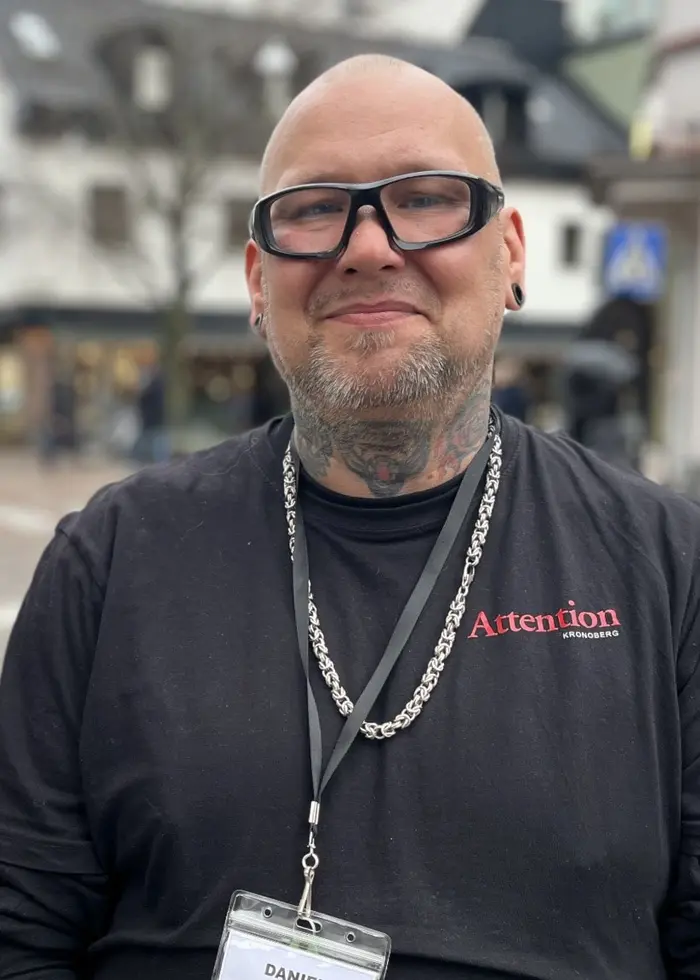 En person står på en gågata omgiven av butiker och snö på marken. Personen bär en mörk tröja med ett namnkort runt halsen. Det är molnigt och träd utan löv syns i bakgrunden.