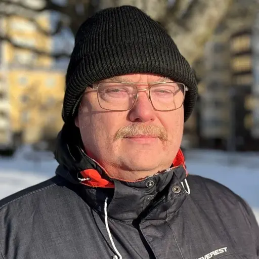 En person klädd i en mörk vinterjacka står utomhus på en snötäckt gångväg med flerfamiljshus i bakgrunden. Solen skiner och skuggor faller på marken.