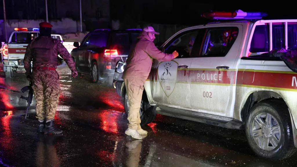 Syriska demokratiska styrkor (SDF) ses vid en väg med flera fordon, inklusive en militärpolisbil, på natten. Reflektioner av ljus från fordonen syns på den våta asfalten.