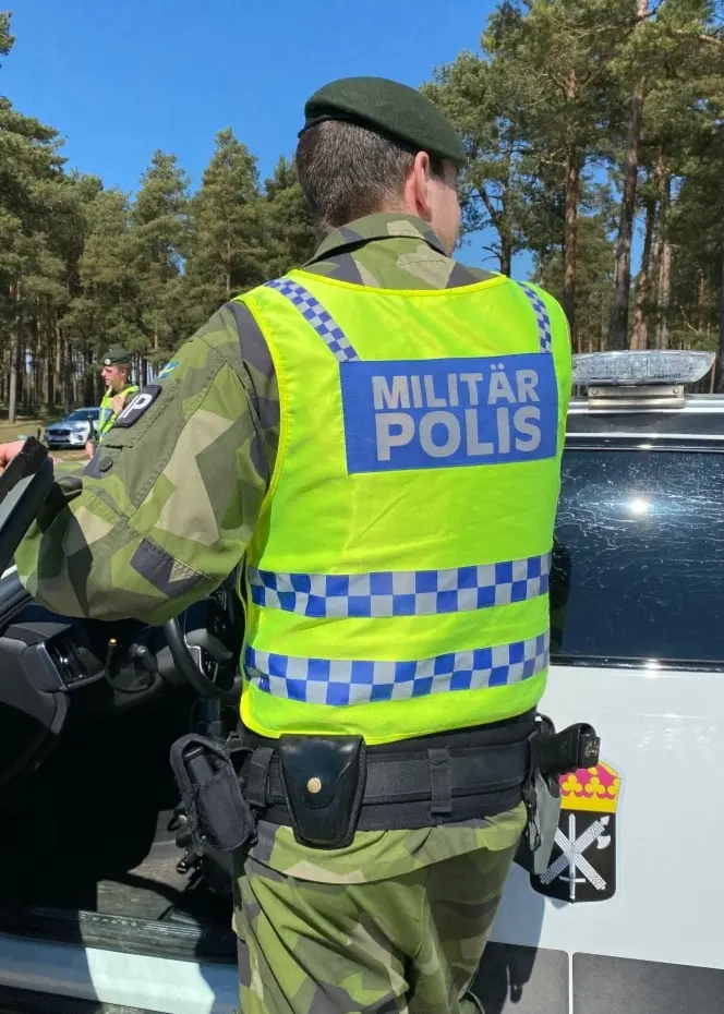 En militärpolis i uniform och reflexväst står bredvid en vit militärpolisbil med tydlig märkning, parkerad på en grusväg. Skog syns i bakgrunden under en solig dag.