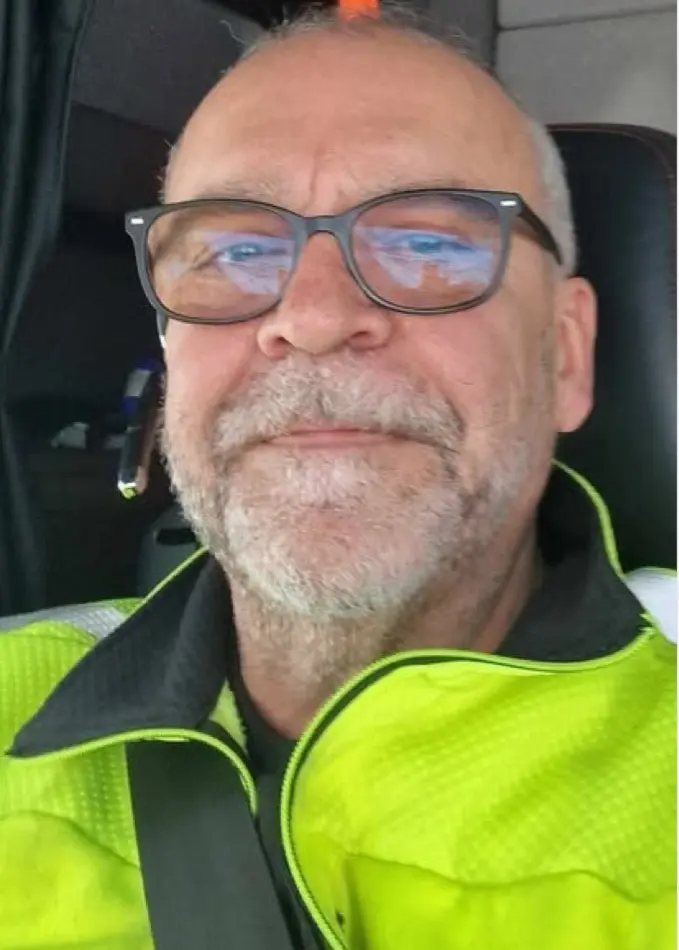 En plogbil med röd plogblad kör på en snötäckt väg och skapar stora snömoln. Bilen har varningsljus på taket. I bakgrunden syns vinterlandskap med snöklädda träd. Till vänster bild på Gunnar Gustavsson, sittandes i lastbilen iklädd varseljacka.