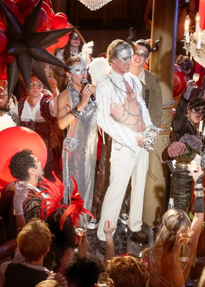 En festlig scen med många människor i eleganta kläder. En person i vit kostym står i centrum omgiven av glamorös dekoration, röda ballonger och en stor kristallkrona i taket.