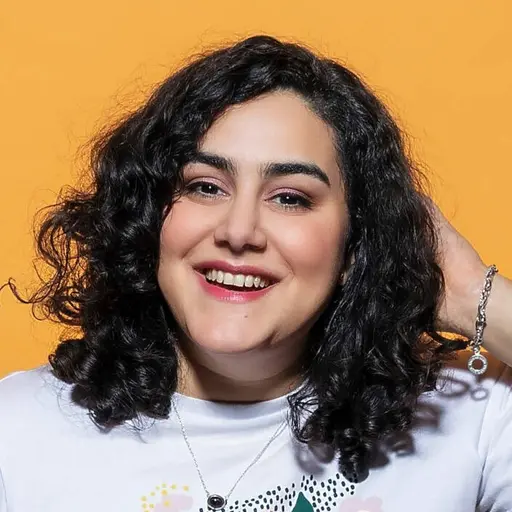 Tina står framför en orange bakgrund. Personen bär en vit T-shirt med ett färgglatt tryck av en hand och grafiska detaljer.