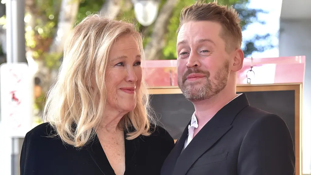 Två personer står tillsammans utomhus framför en skylt på Hollywood Walk of Fame. Ena personen bär en svart klänning och den andra en mörk kostym. Träd och lampor syns i bakgrunden.