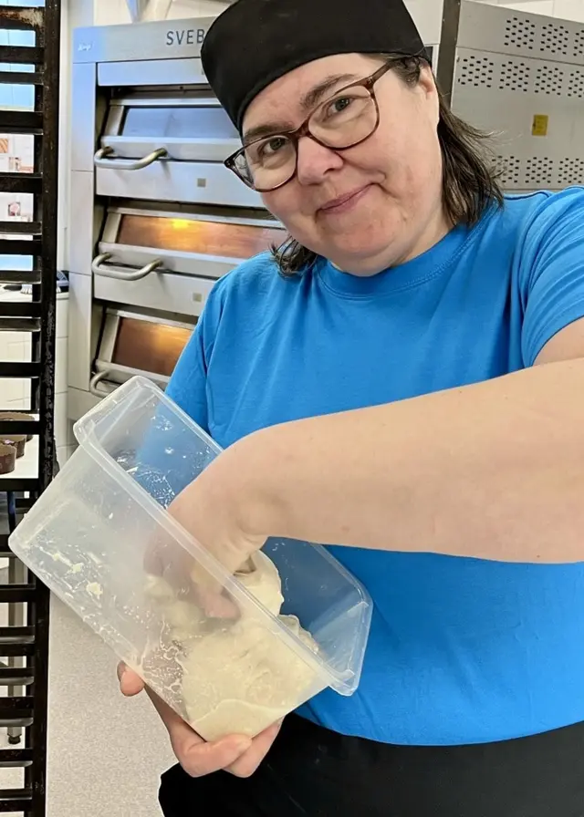 Bagare Camilla Andersson i blå tröja arbetar med att baka beredskapsbröd i ett bageri med hyllor fyllda av bröd i bakgrunden.