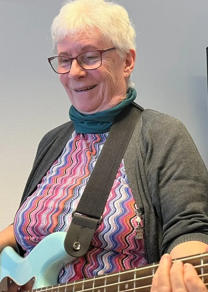 En person spelar elbas i ett rum med en högtalare i bakgrunden. Personen bär en färgglad klänning och en grå kofta. Elbasen är ljusblå med metallfärgade detaljer.