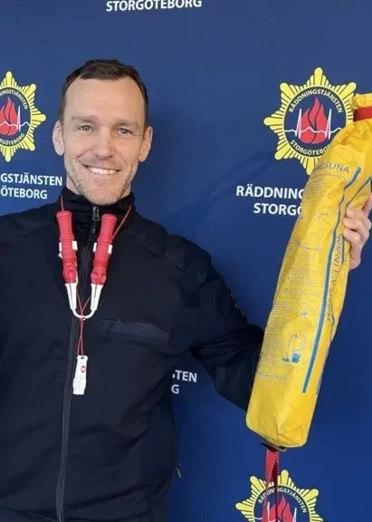 En person i uniform håller i två föremål, en svart flytväst och en gul kastlina, framför en blå bakgrund med Räddningstjänsten Storgöteborgs logotyp och text.