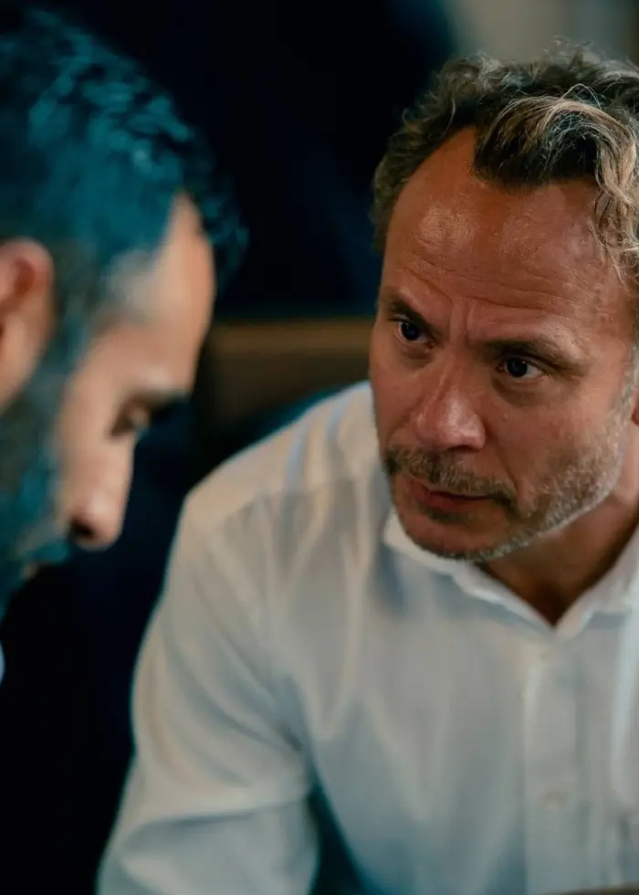 Två personer i vita skjortor sitter mittemot varandra och samtalar i en inomhusmiljö med dunkel belysning.