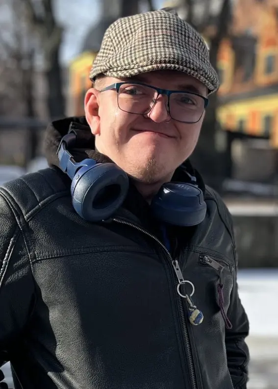 Person står utomhus i vintermiljö med snö på marken och orangea hus i bakgrunden. Personen bär en mörk jacka och hörlurar runt halsen. Solen skiner och träden är utan löv.