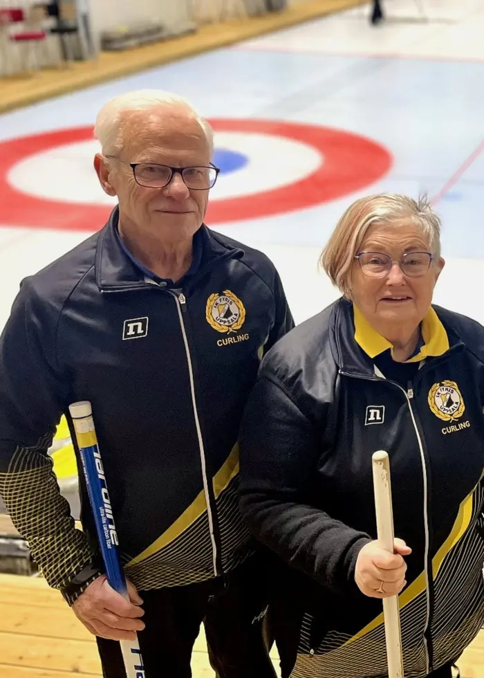 Fyra personer står med curlingklubbor på en curlingbana inomhus. Curlingstenen och isbanan med måltavla syns tydligt i bakgrunden.