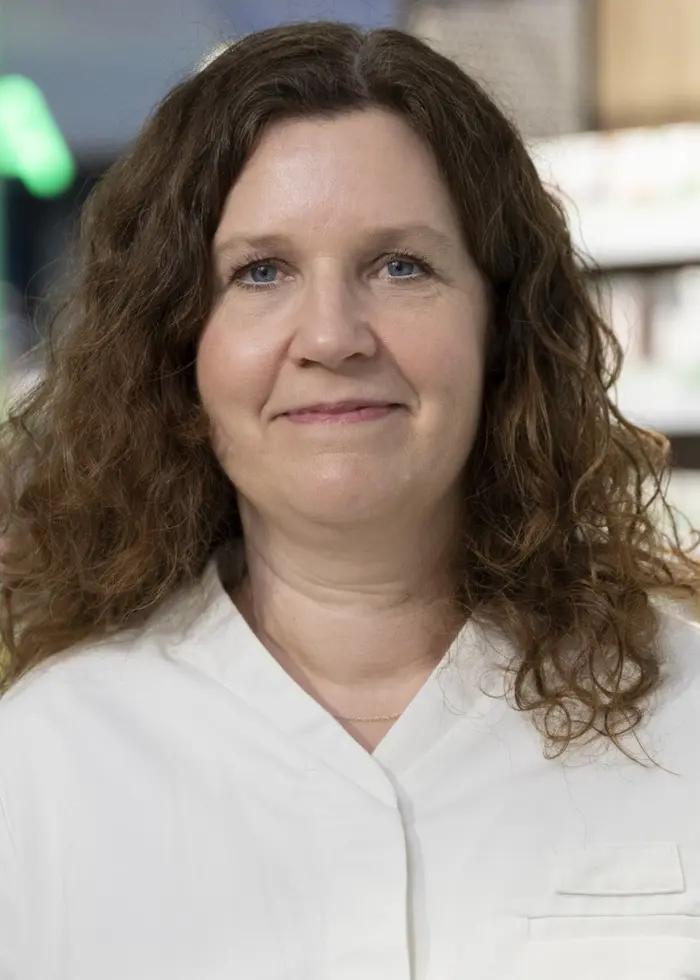 En apotekare med lockigt hår iklädd vit arbetsrock står framför en hylla fylld med medicinprodukter på ett apotek. I bakgrunden syns en oskarp skylt och butiksmiljö.