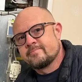 En person iförd en mörk jacka står vid en värmepanna och pekar på en del av installationen. I bakgrunden syns rör och teknisk utrustning monterad på väggen.