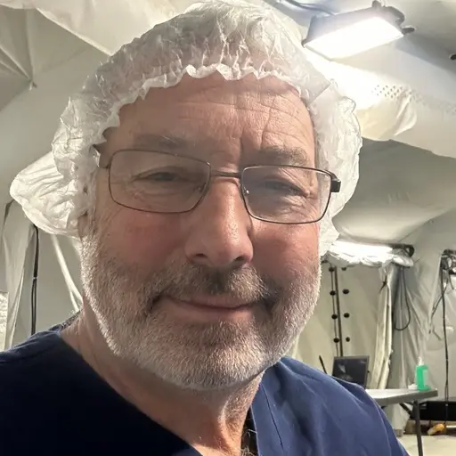 En person i blå sjukhuskläder och skyddsmössa befinner sig i ett tältliknande medicinskt område. I bakgrunden syns medicinsk utrustning och personal i arbete.