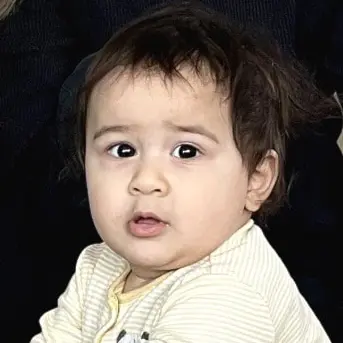 En familj sitter på ett vardagsrumsgolv framför en soffa. Föräldrarna sitter bakom sitt barn som leker med en grön plaststol. Ett blått leksaksdjur och en matta syns i förgrunden.