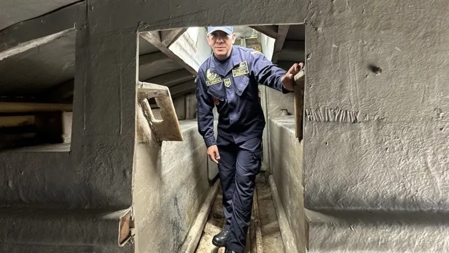 En person i uniform navigerar genom en trång passage inne i en ubåt eller ett liknande fartyg. Omgivningen består av gråa metallväggar och en smal gång.